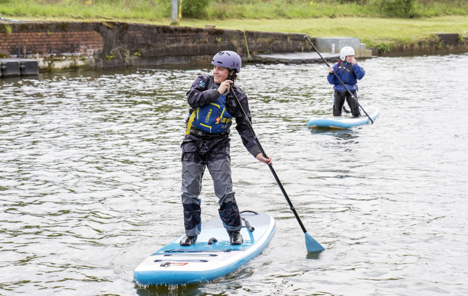 Two people stand-up paddleboarding on a calm canal, both wearing helmets and buoyancy aids.