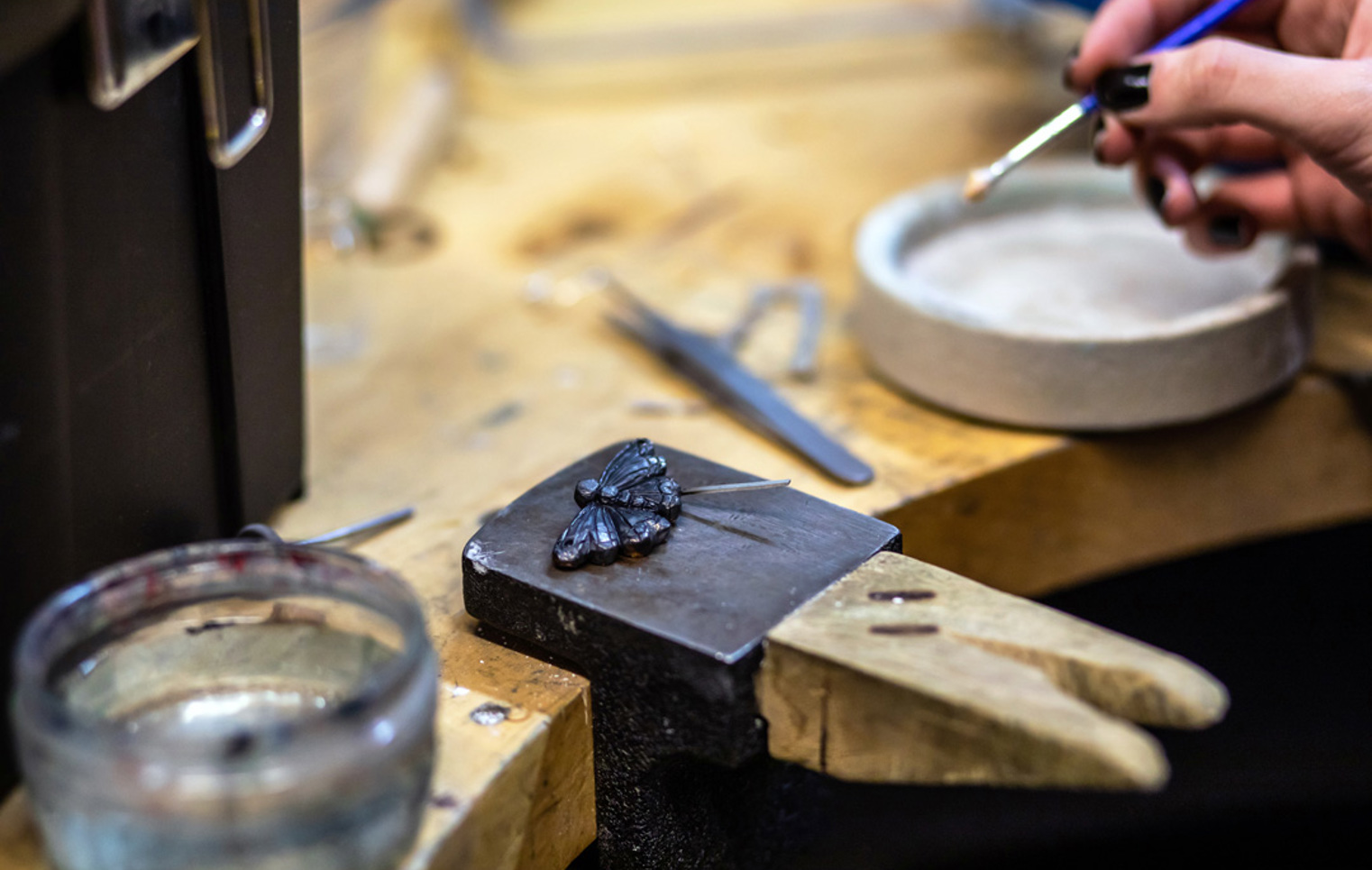 A close-up of a dark metal butterfly-shaped jewellery piece on a metal block, with an artist’s hand holding a fine brush to apply detail work.