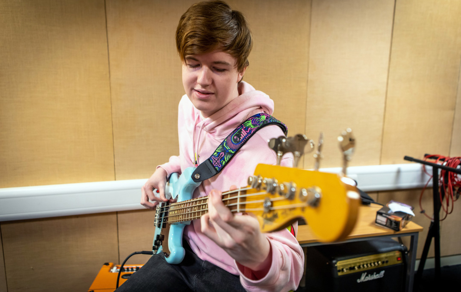 A young student in a pink hoodie playing a blue bass guitar in a music practice room, focusing on their instrument.