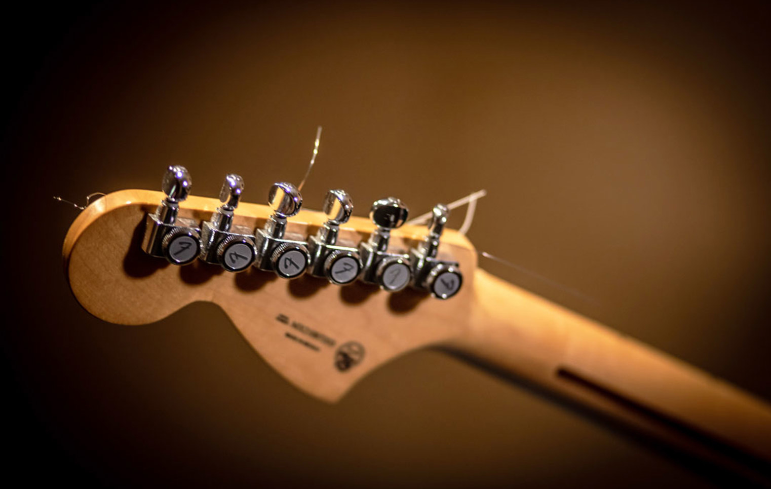 A wooden guitar headstock with chrome tuning pegs and loose strings, set against a blurred background.