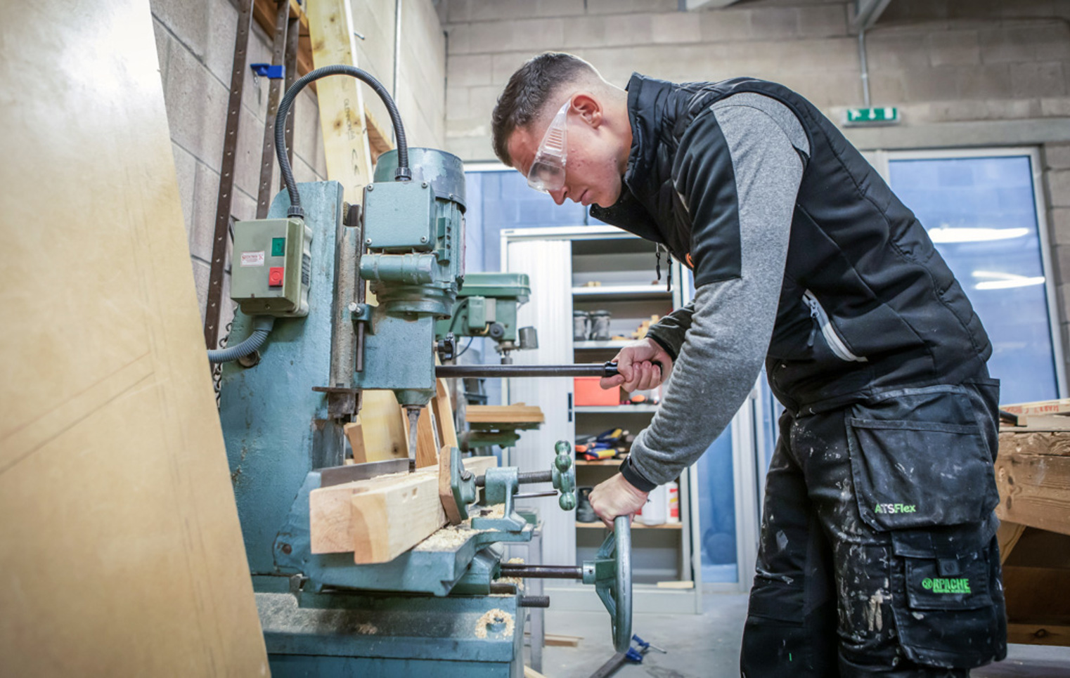 A joinery student wearing safety glasses, operating woodworking machinery to shape timber in a college workshop.