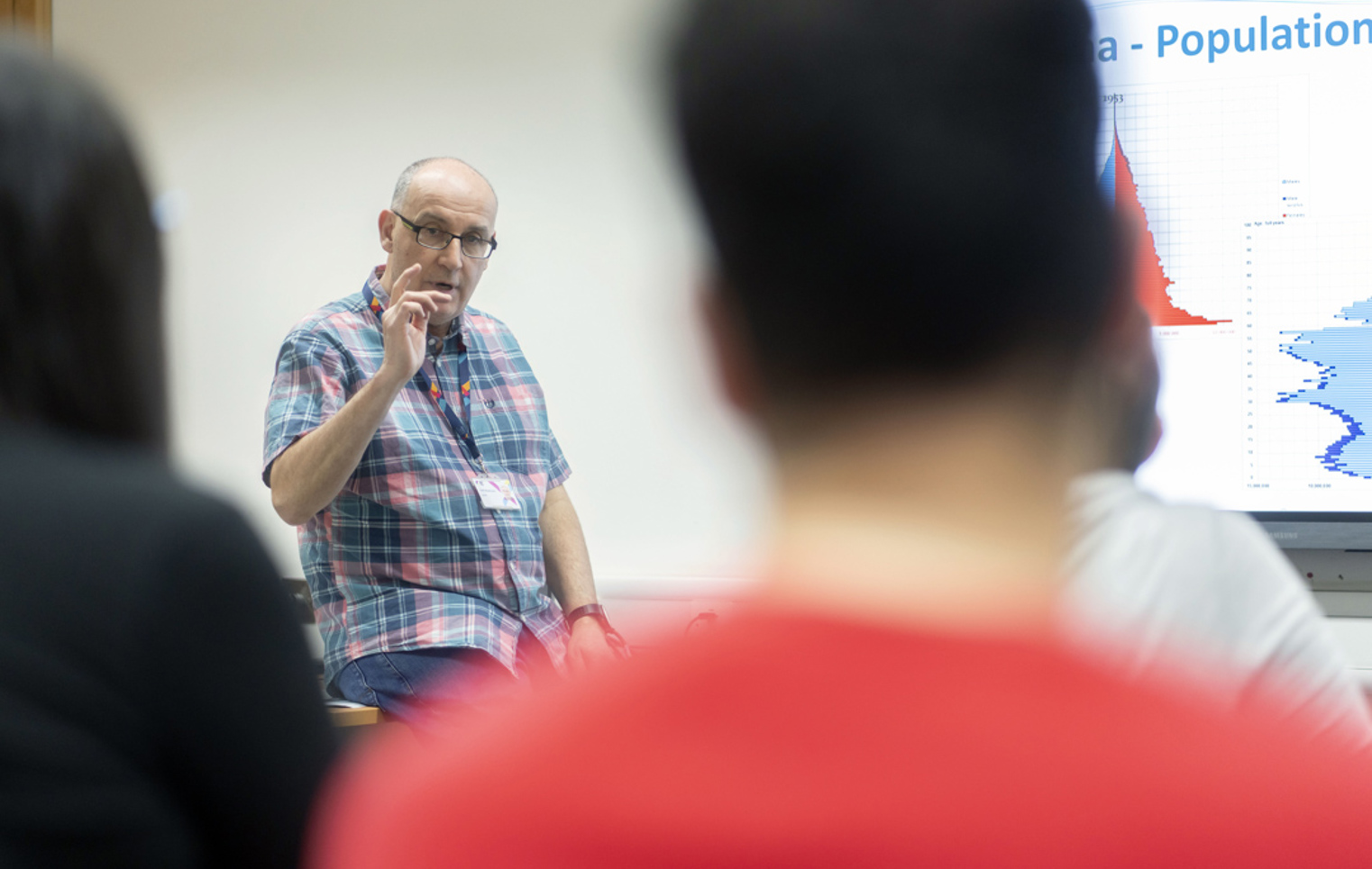 A lecturer engages with students in a classroom at Glasgow Kelvin College, with population graphs on screen.