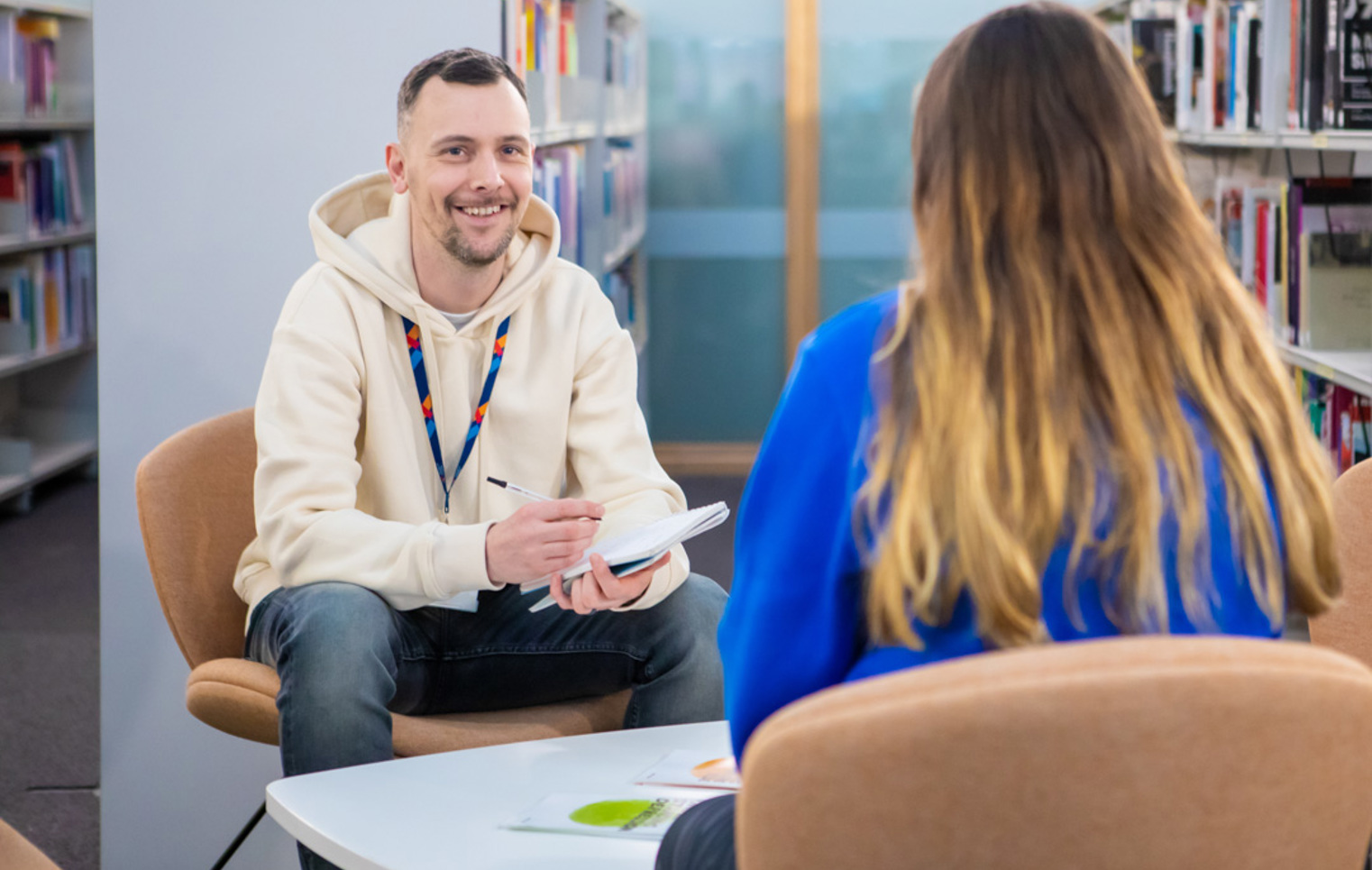 Glasgow Kelvin College staff member in a cream hoodie speaking with a student in a library setting, both seated and engaged in conversation with a notepad.