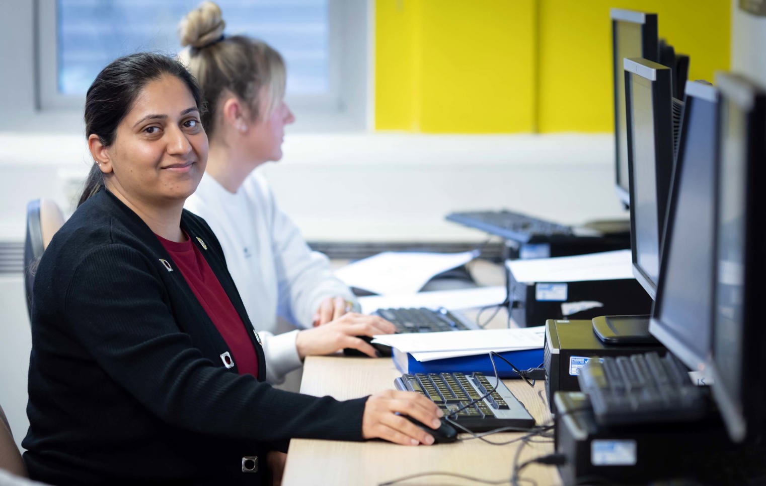 Student in a black cardigan sitting at a computer workstation, smiling while working on a task in a business and admin class.