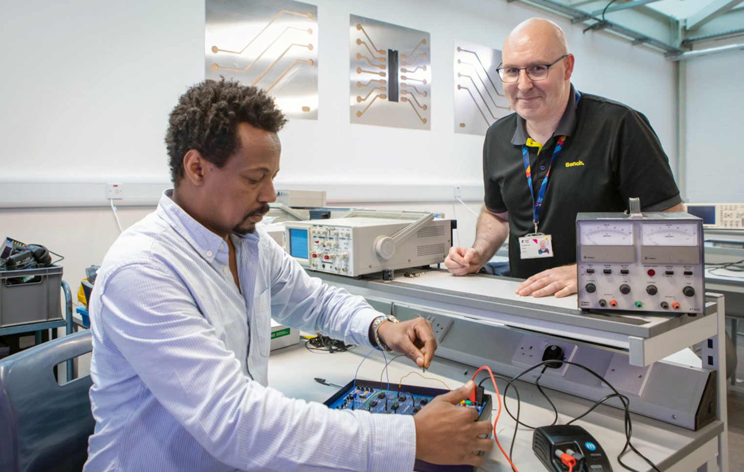 A lecturer and a student in an electronics lab working on a circuit board experiment. The lecturer is smiling while overseeing the student’s work.