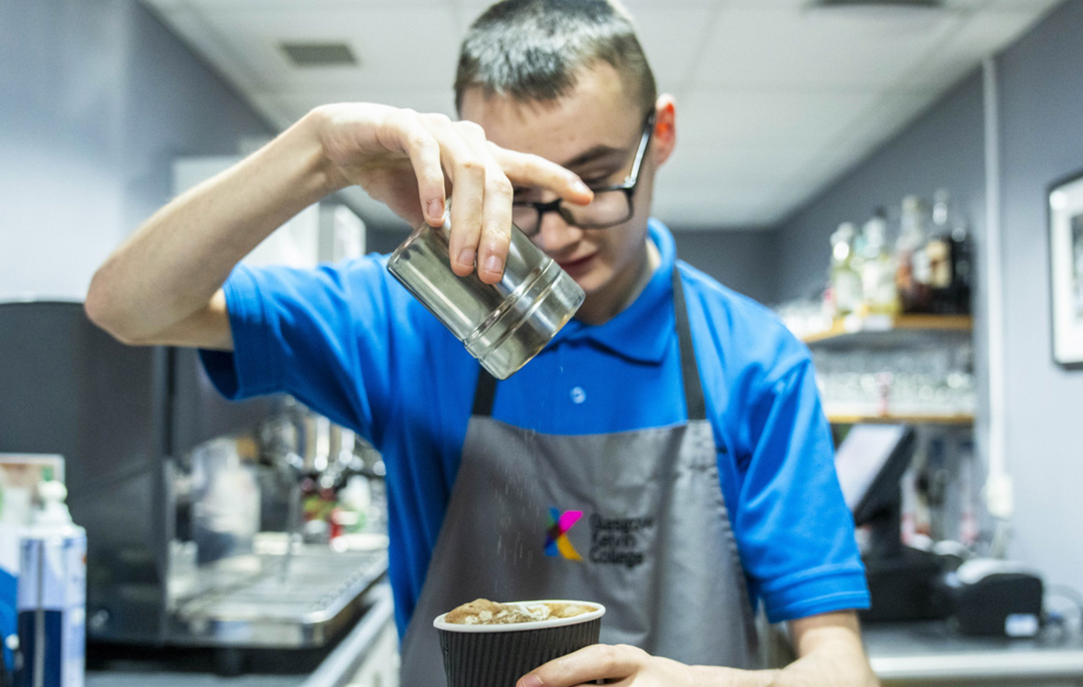 A young barista in a blue uniform sprinkles cocoa powder over a freshly prepared cup of coffee in a café setting.
