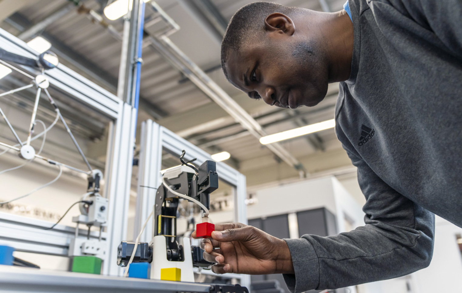Student adjusting a robotic arm to pick up a red block, focusing intently on precision in a mechatronics workshop.