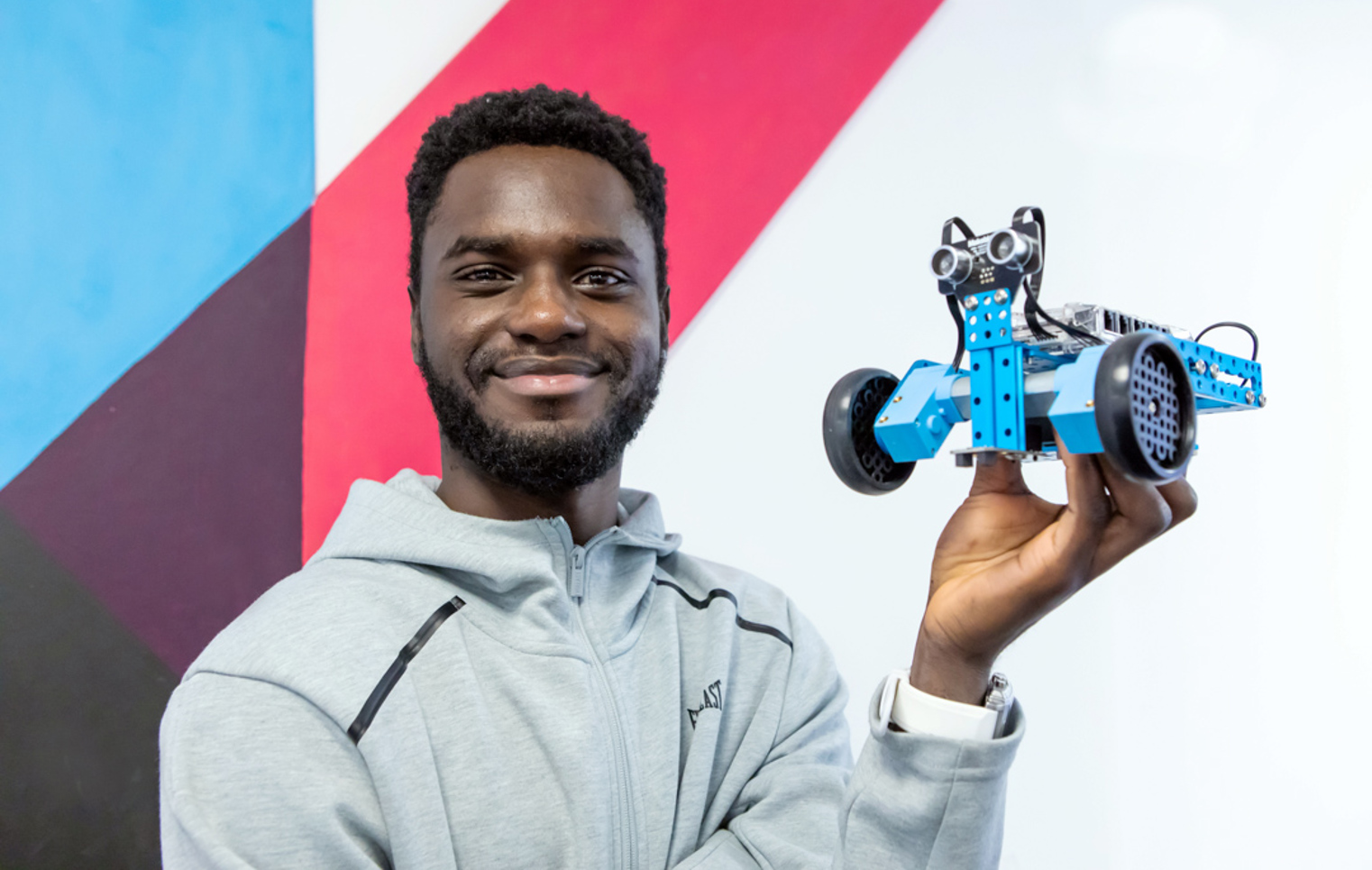 Student in a grey hoodie holding up a blue robotic vehicle, smiling against a colourful geometric wall.