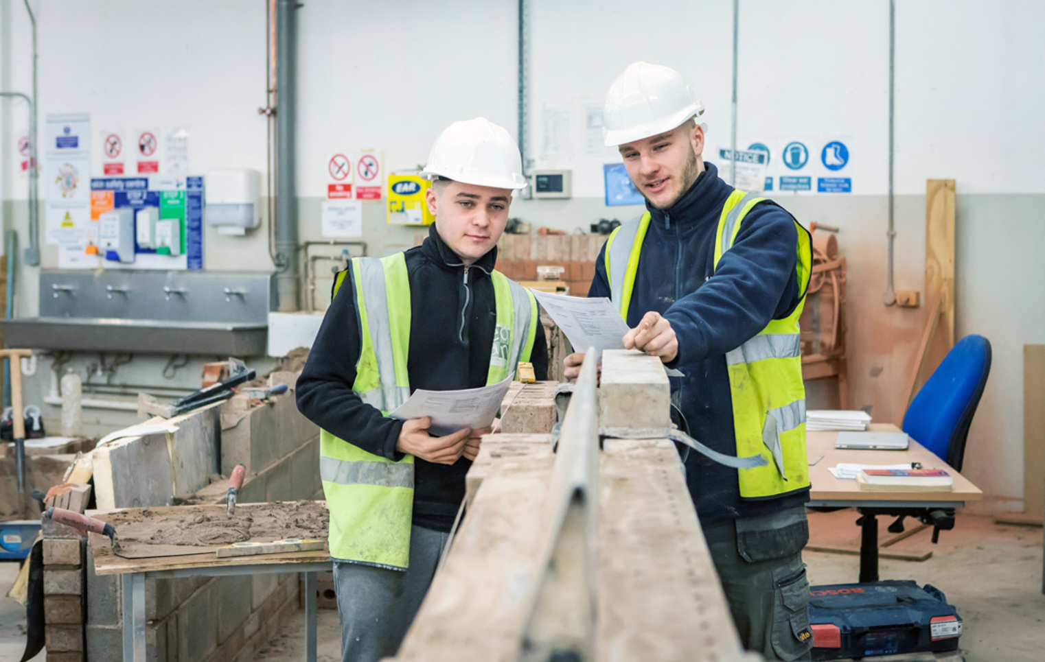 Two brickwork students in safety gear reviewing plans together, standing beside a brick wall and discussing their next steps in a practical training workshop.
