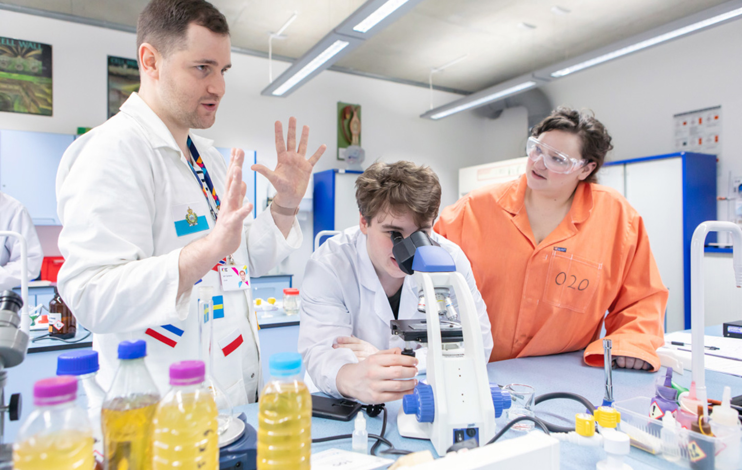 Science lecturer guiding a student on microscope use with classmates observing in the lab at Glasgow Kelvin College