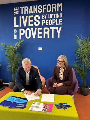 Partnership agreement being signed beneath a “We Transform Lives by Lifting People Out of Poverty” message wall.