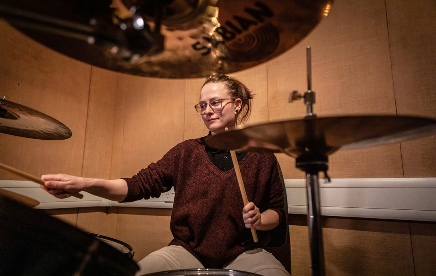 A young drummer with glasses and a burgundy sweater, playing a drum kit surrounded by cymbals.