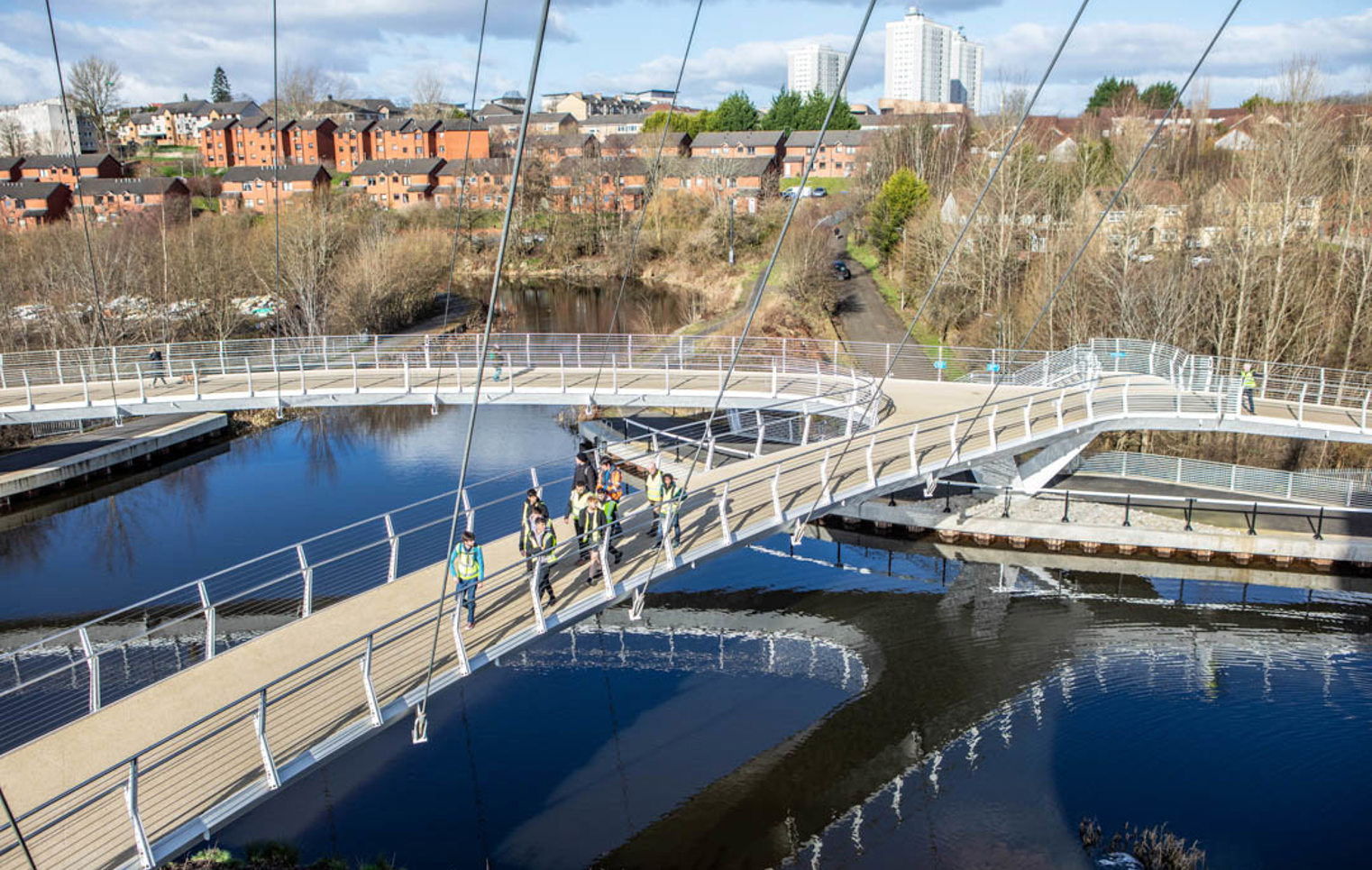 Overhead view of students and lecturers walking across the curved paths of the Stockingfield Bridge, with the canal reflecting the structure and surrounding landscape.