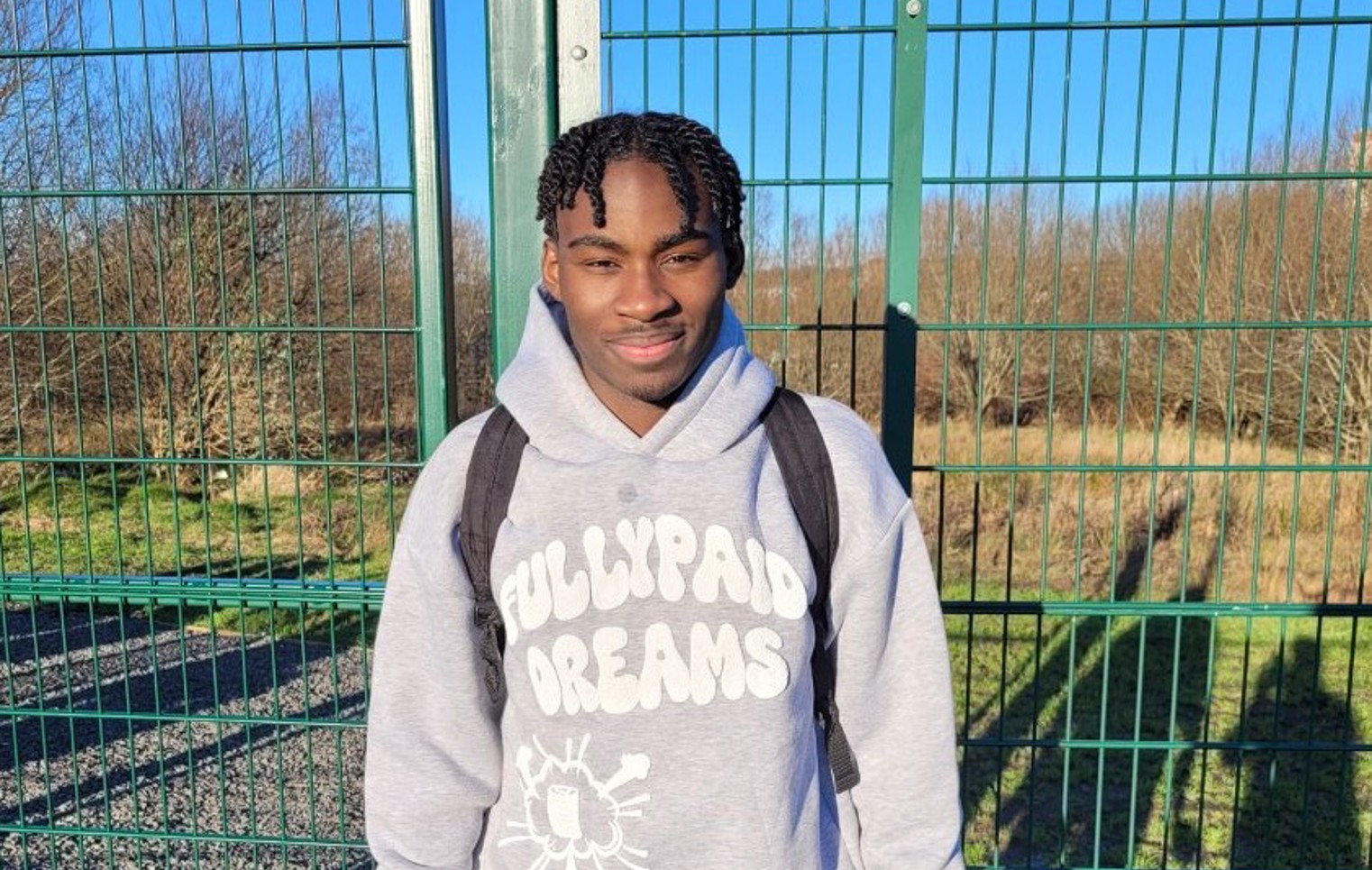 Glasgow Kelvin College student Tuacenio Santos standing outside on a sunny day in sportswear, holding headphones and a water bottle, smiling at the camera.