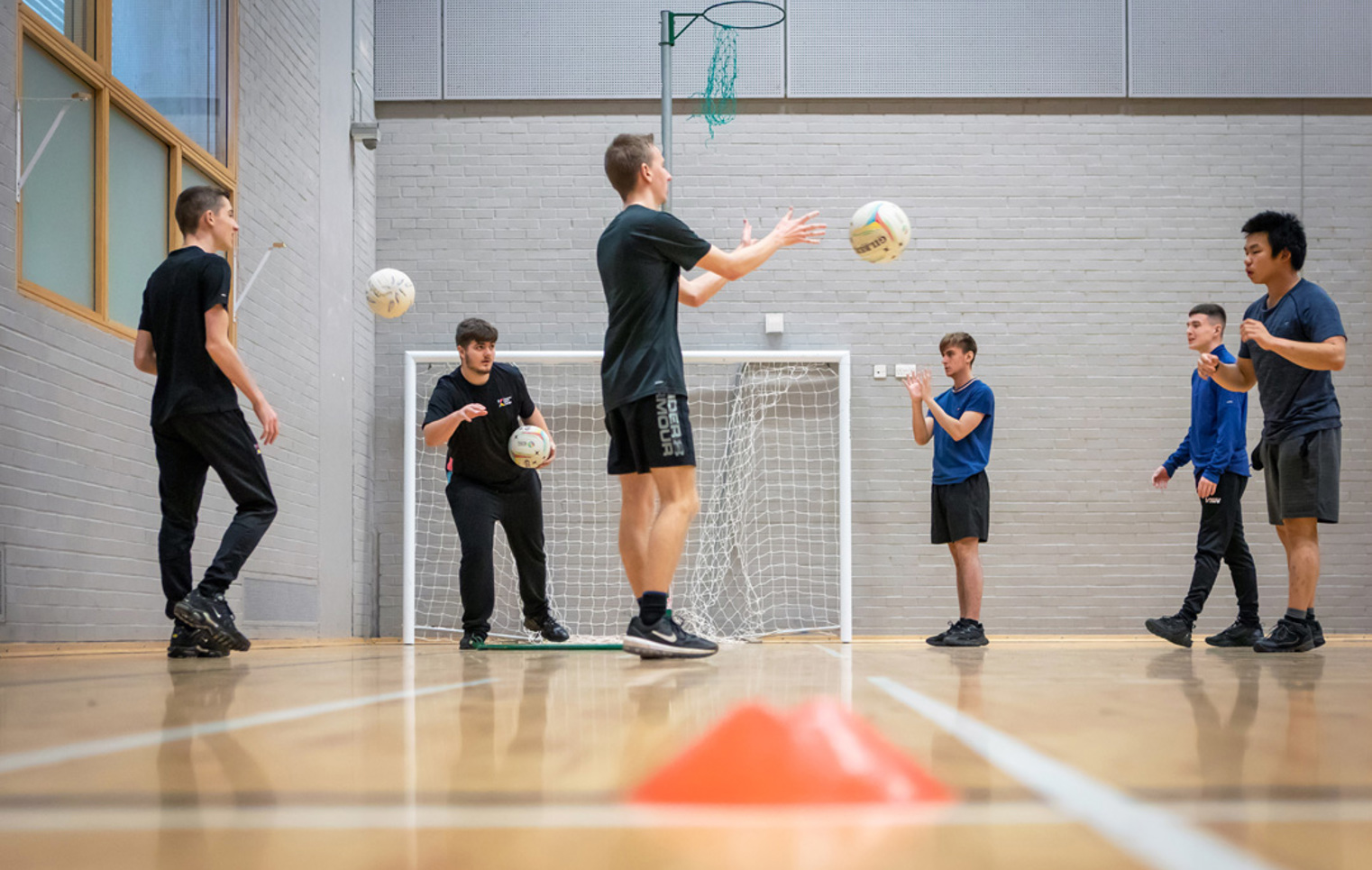 Students in a games hall tossing netballs in a circle as part of a group activity.