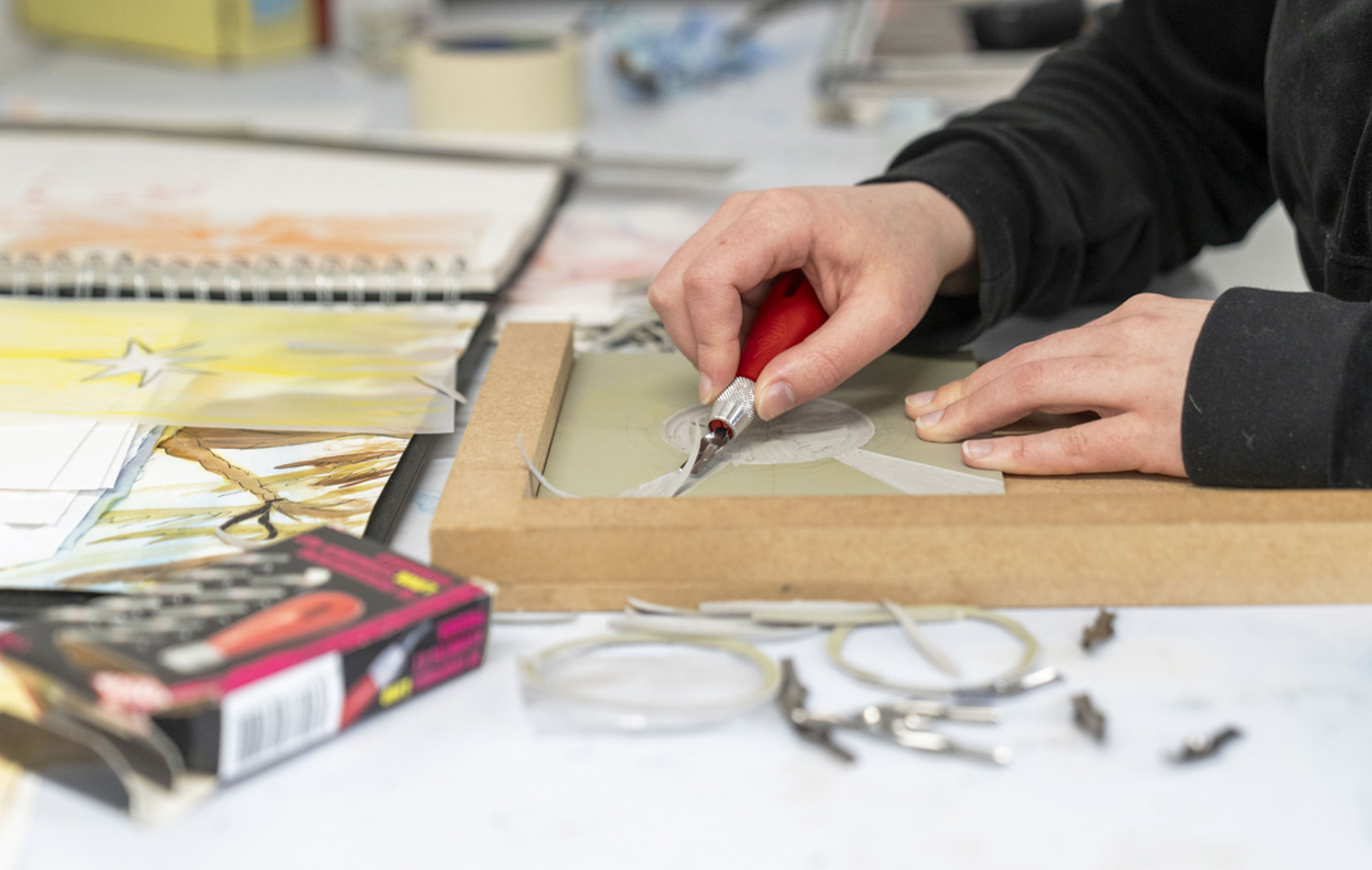 Close-up of hands carving a design into a printmaking block on a desk, surrounded by sketches and art supplies