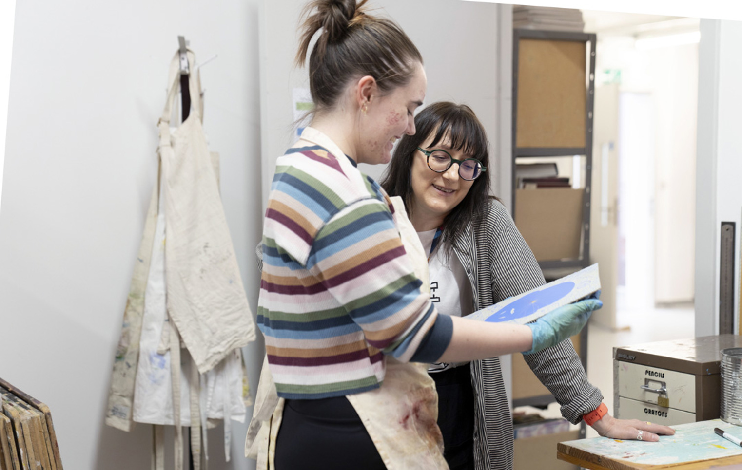 Two people in an art studio discussing a blue printmaking plate, with aprons and art supplies in the background.