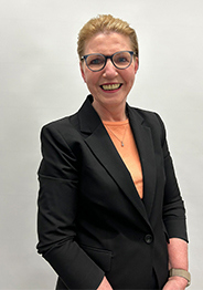 Lesley Quinn, a woman with tied-back hair wearing glasses, a black blazer, and an orange top, smiling against a plain light background