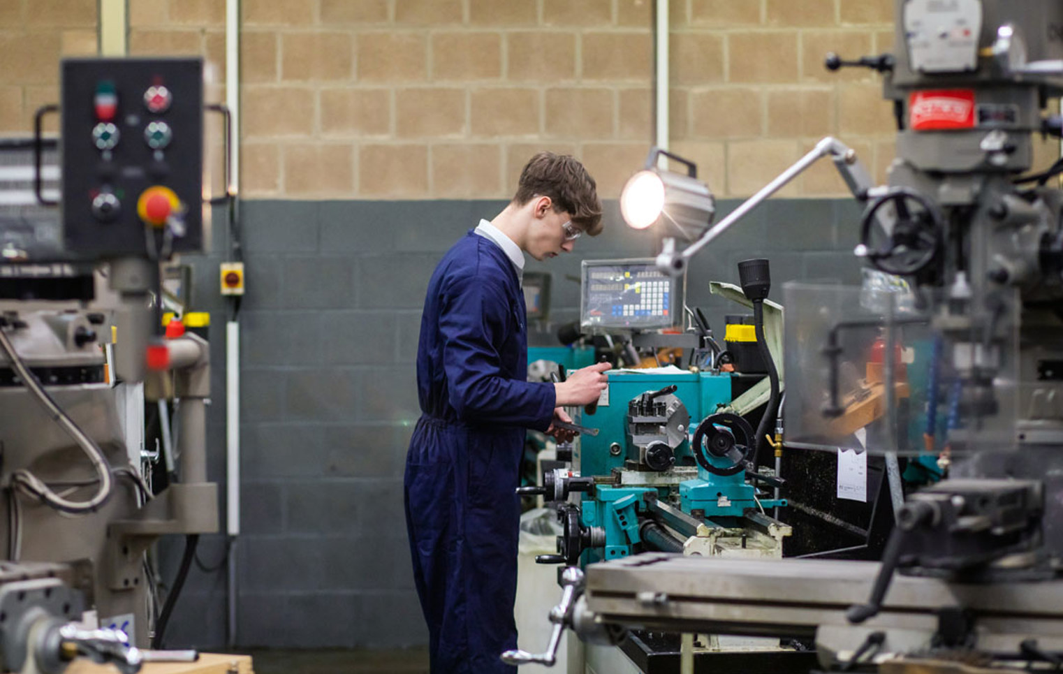 A student in navy-blue overalls operating a lathe machine in a mechanical engineering workshop, focused on precision machining.