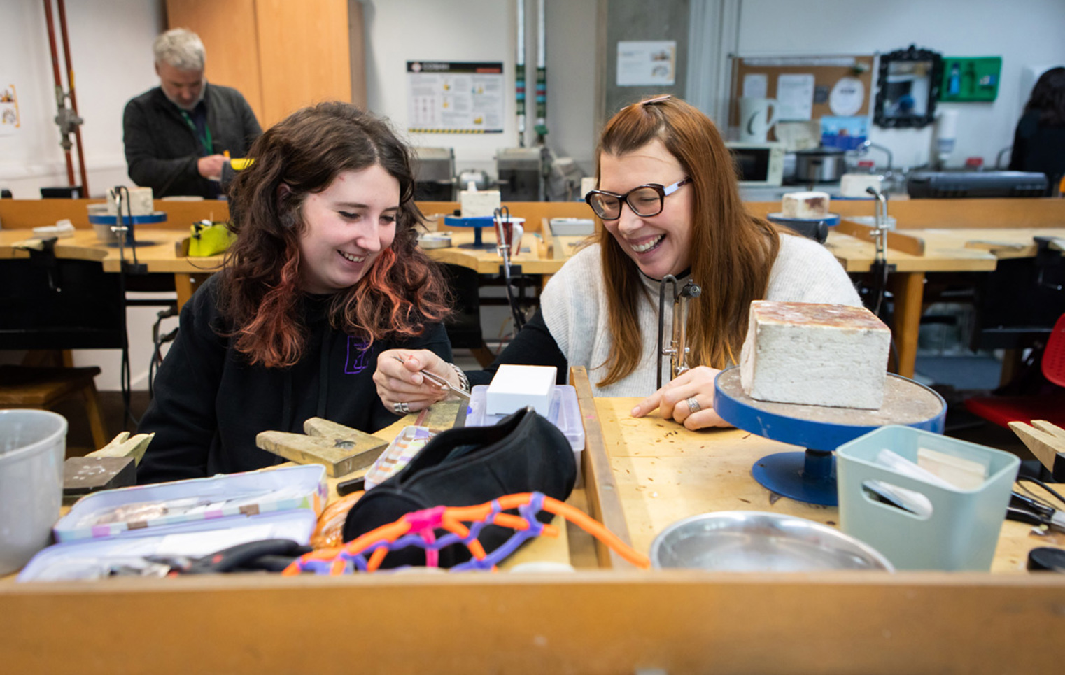 Two women in a jewellery workshop laughing and working together at a wooden workbench, with various tools and equipment scattered around.