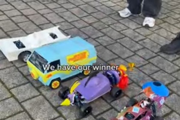 A group of people standing around three colorful, custom-built model cars at the finish line of the Annual Kelvin CAD Car Race. The winning car, a bright blue and yellow van, is in the foreground with a "Winner" sticker, while the other two cars, one purple and one red, are beside it on a paved surface.