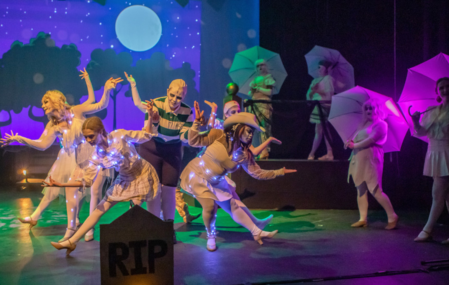 Musical theatre students performing a stylised ensemble dance on stage, with umbrellas, illuminated costumes, and a moonlit backdrop.