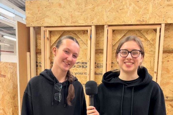 Two women construction students stand smiling in a workshop, both wearing black hoodies and work trousers. One holds a microphone as they talk about the Women in Construction course.