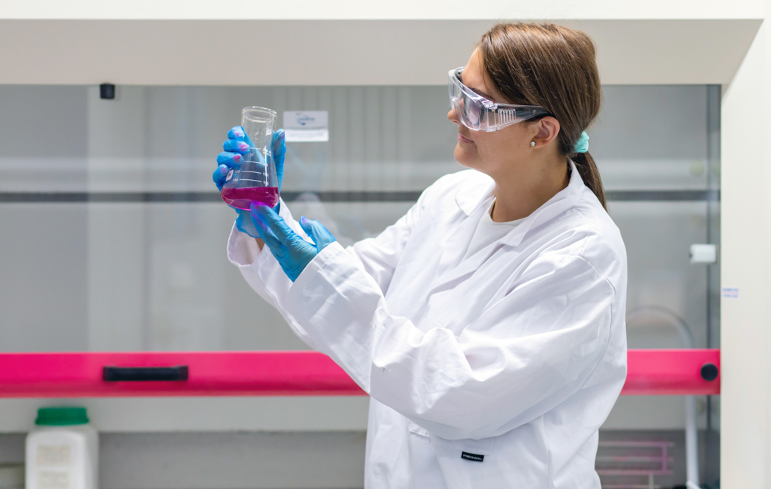 Student in a lab coat studying a beaker of pink liquid under bright lighting in a science lab.