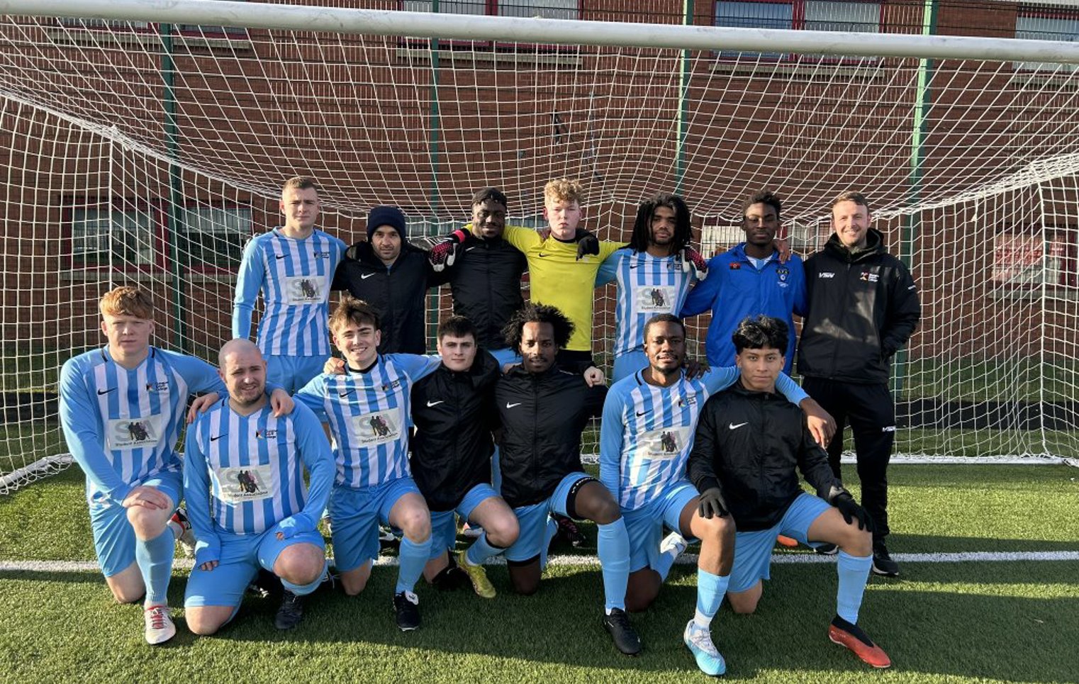 Glasgow Kelvin College men's football team posing together in front of a goal on the pitch, wearing blue and white kits.