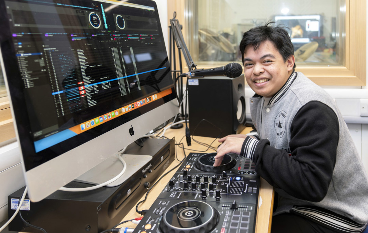 Young man smiling while using DJ equipment and a microphone, with a computer screen displaying music software.