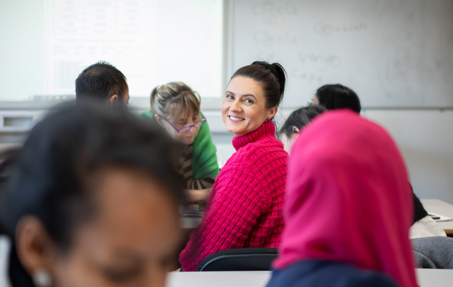  A woman in a bright pink sweater smiles at the camera while sitting in a classroom with other students working in the background.