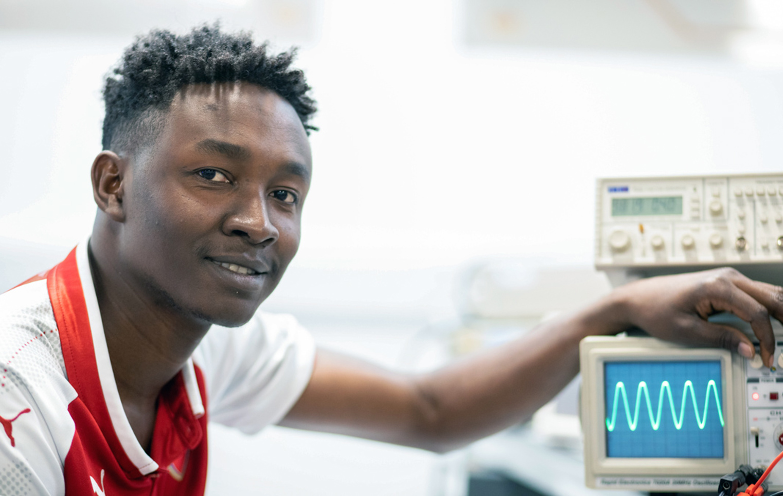 Close-up of a student in a red football jersey adjusting an oscilloscope, displaying a waveform on the screen.