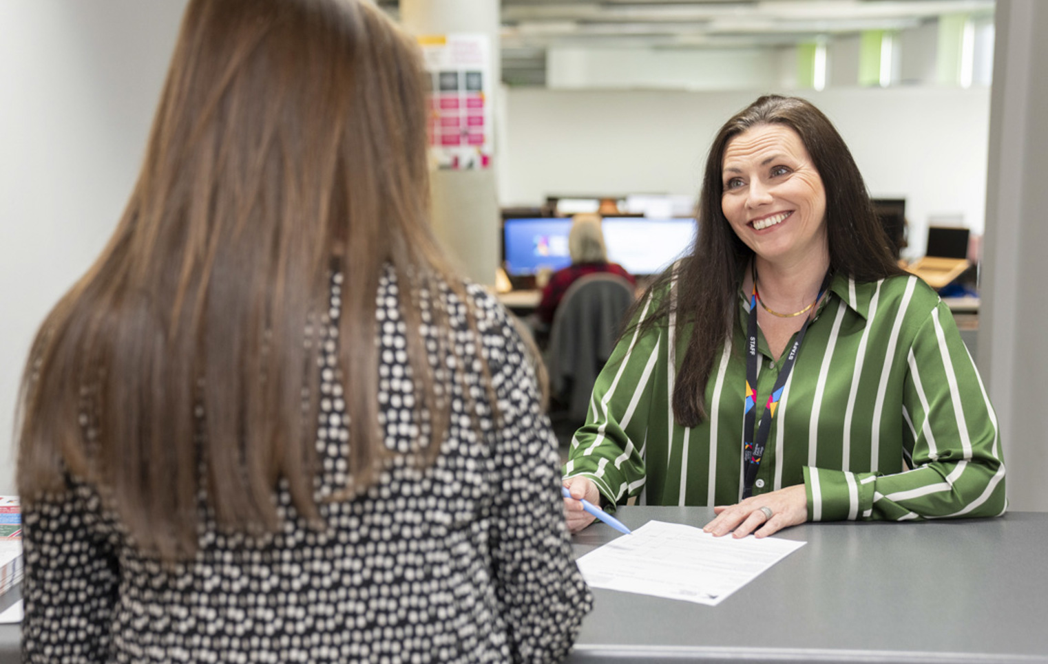 Finance staff member at Glasgow Kelvin College smiling while assisting a student at the service desk, with paperwork on the counter and computers in the background.